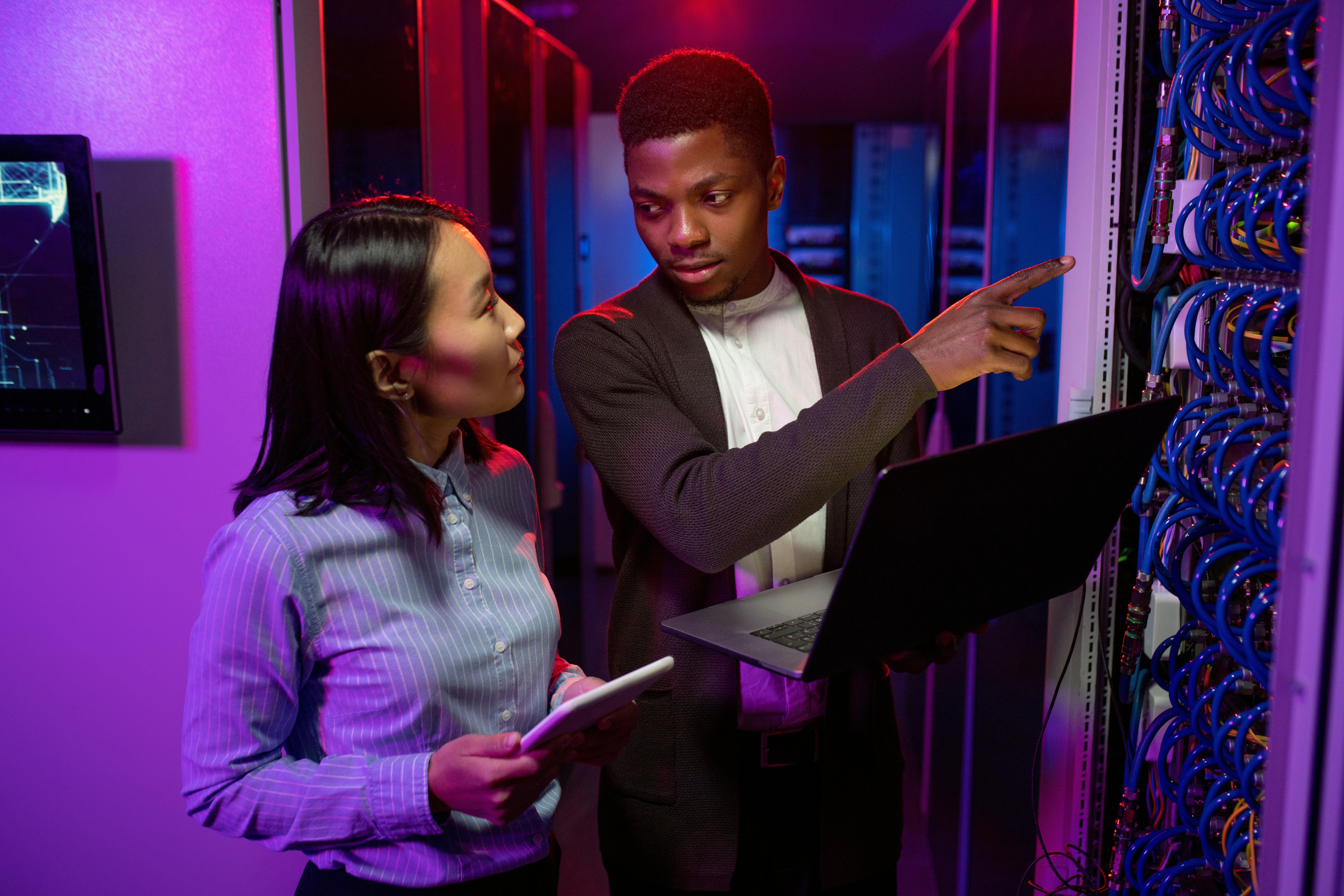 Young Afro-American technician pointing at cables while explaining server productivity to Asian colleague in data center illuminated with neon light photo